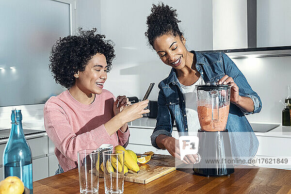 Smiling woman photographing friend blending juice in kitchen at home