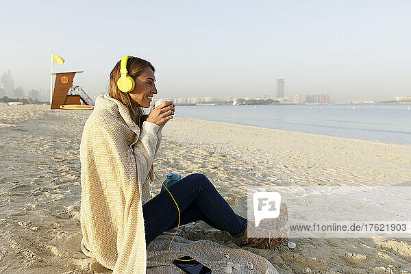 Happy woman holding tea cup listening music through wireless headphones sitting at beach