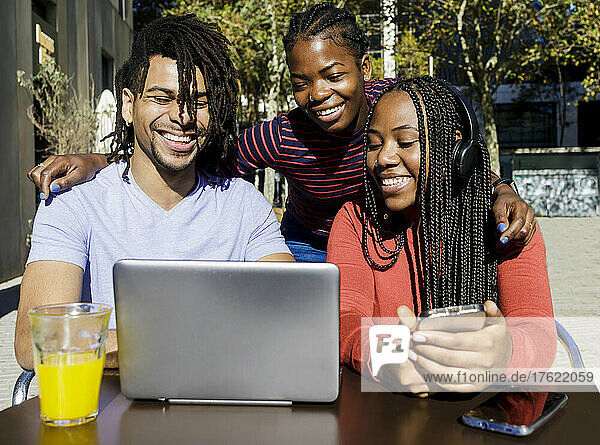 Happy young man and women sharing laptop at sidewalk cafe on sunny day
