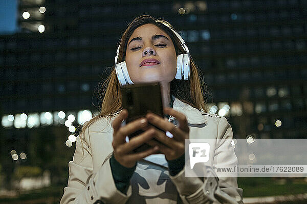 Woman with eyes closed holding mobile phone listening music through headphones at night