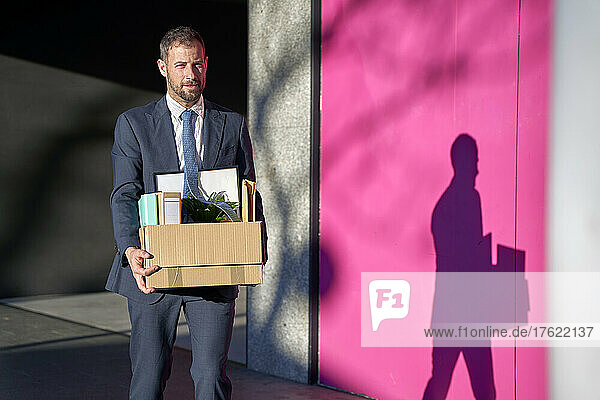 Businessman carrying box of personal belongings by wall on footpath