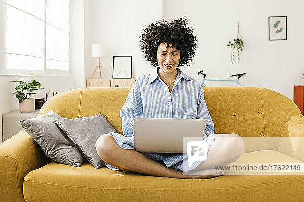 Smiling young woman using laptop sitting cross-legged on sofa at home