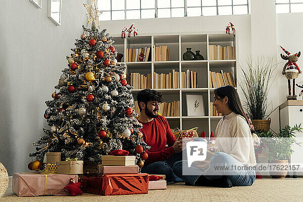 Happy couple holding gift box sitting by Christmas tree at home