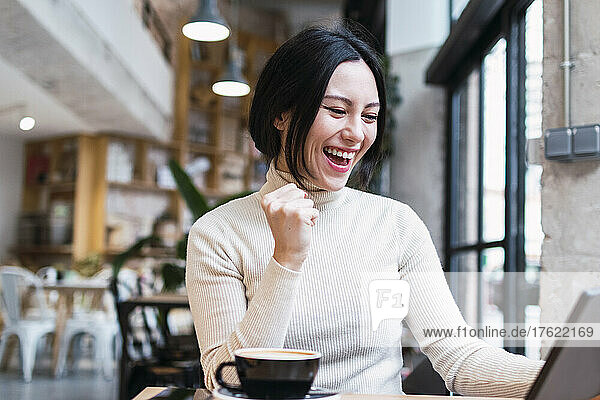 Happy woman holding tablet PC gesturing fist sitting at cafe