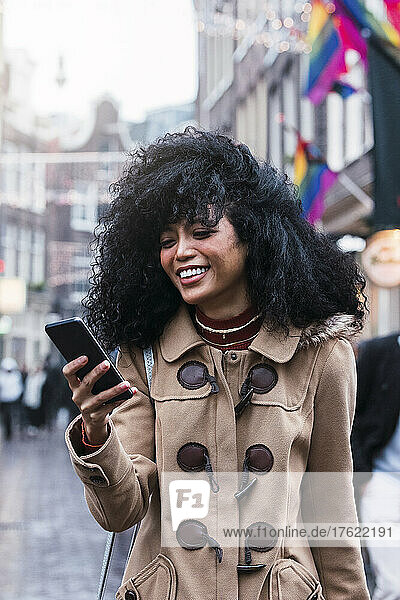 Happy young woman with tousled hair surfing net through smart phone in city