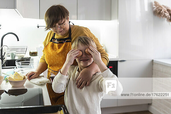 Smiling boy covering eyes with eggs standing by grandmother in kitchen