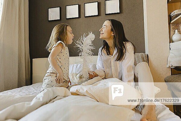 Playful girl blowing on feather held by woman siting with eyes closed on bed at home