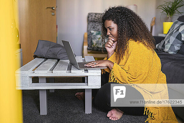 Young curly haired woman sitting cross-legged using laptop at coffee table in living room