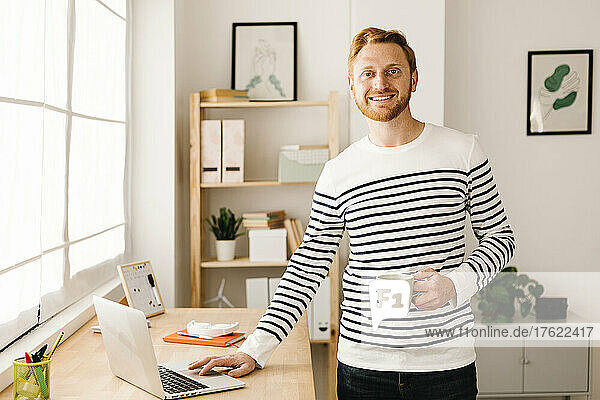 Smiling young man holding coffee cup standing by laptop at home