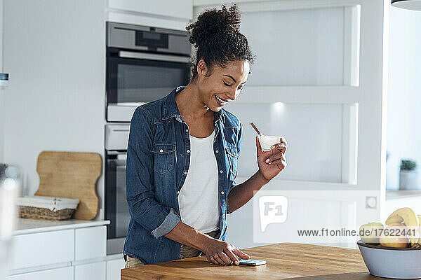 Smiling woman holding bowl using mobile phone in kitchen