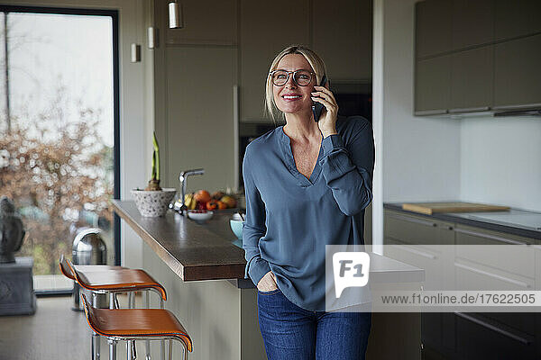 Lächelnde blonde Frau  die zu Hause mit der Hand in der Tasche am Küchentisch steht und mit ihrem Smartphone spricht