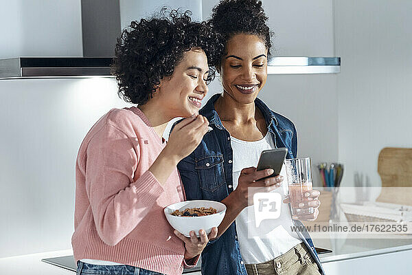 Smiling women sharing smart phone in kitchen at home