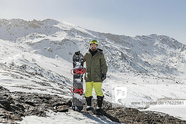 Smiling young man standing with snowboard on rock against snowcapped mountain