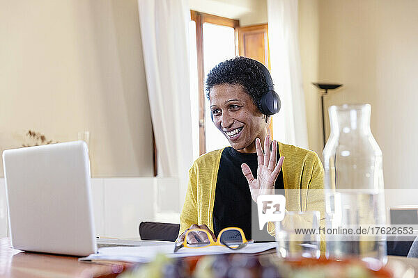 Smiling businesswoman with headphones waving on video call through laptop at home