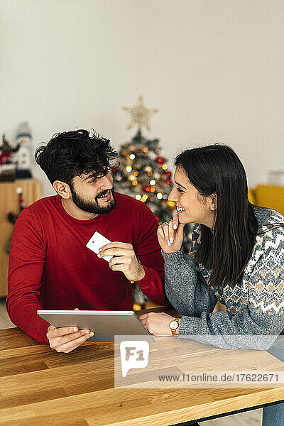 Lächelnder Mann hält Tablet-PC und Kreditkarte in der Hand und blickt seine Freundin zu Hause mit der Hand am Kinn an