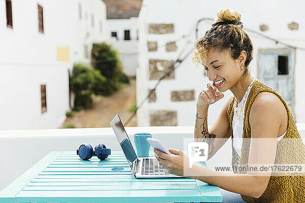 Happy woman using smart phone sitting with laptop at table on rooftop