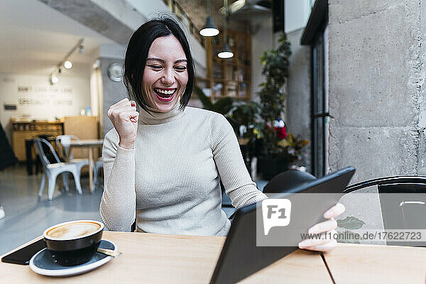 Cheerful woman gesturing fist holding tablet PC sitting in cafe