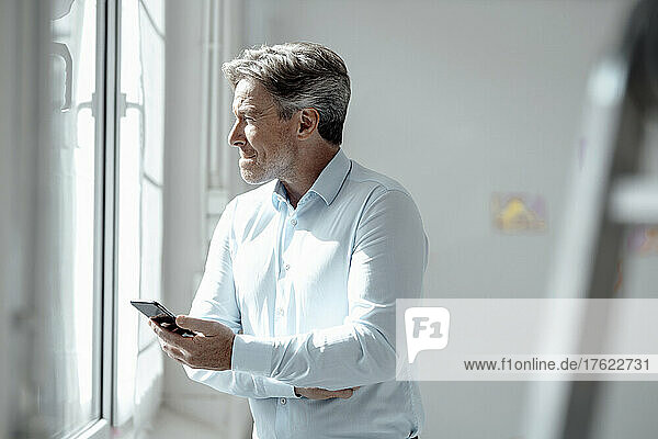Businessman with smart phone looking out through window at office