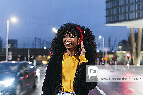 Happy young woman listening music through wireless headphones on street at night