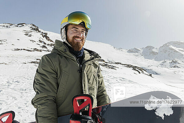 Happy young man wearing ski goggles holding snowboard on sunny day