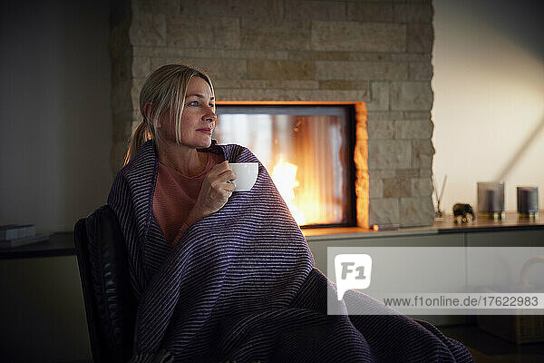 Blond woman wrapped in blanket holding coffee cup sitting at home