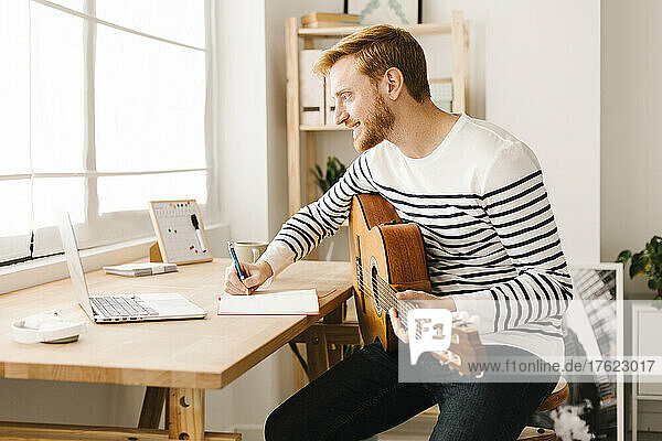 Lächelnder junger Mann mit Gitarre  der zu Hause Musiknoten schreibt und am Laptop lernt