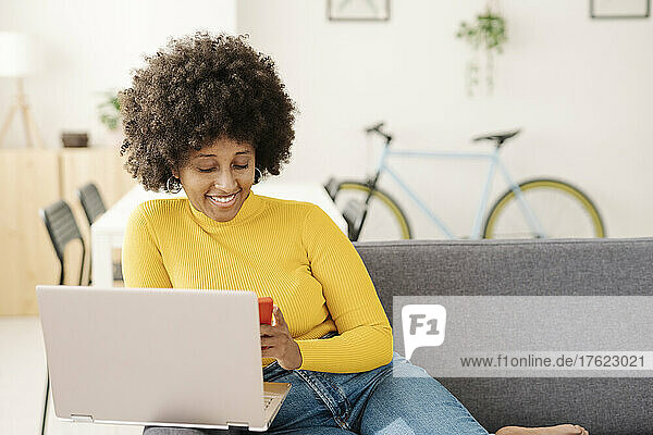 Smiling young woman using smart phone sitting with laptop on sofa in living room