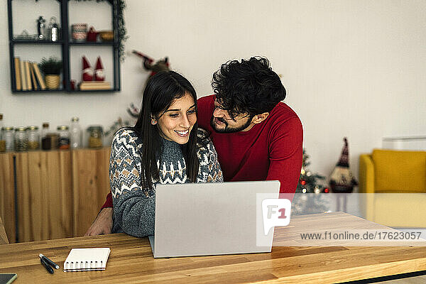 Junger Mann umarmt lächelnde Freundin  die zu Hause Laptop benutzt