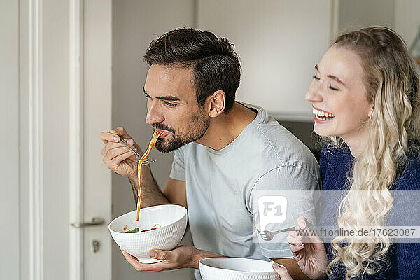 Happy young man eating spaghetti by cheerful girlfriend at home
