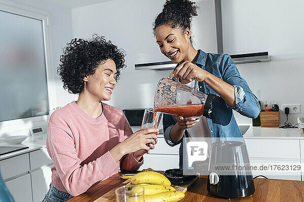 Smiling woman pouring juice for friend into glass in kitchen at home