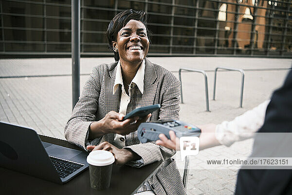 Glückliche Geschäftsfrau  die kontaktlos per Smartphone an die Kellnerin im Straßencafé bezahlt