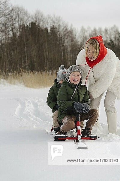 Happy mother pushing sons sitting on toboggan in winter