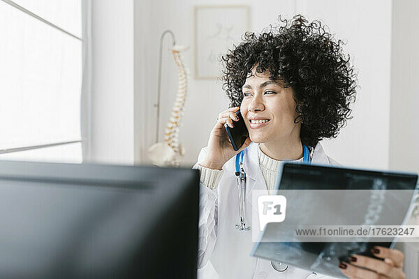 Smiling doctor with X-ray talking on smart phone sitting at medical clinic