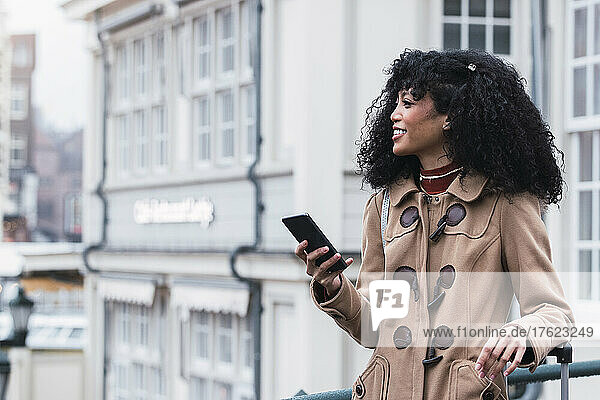 Smiling woman with curly hair holding mobile phone in city