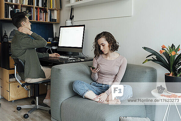 Young man using desktop PC by woman surfing net through smart phone on sofa at home