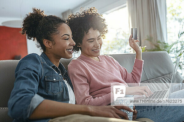 Smiling young woman holding credit card doing online shopping through laptop sitting by friend at home