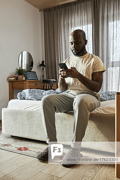 Man using smart phone sitting on bed in morning at home