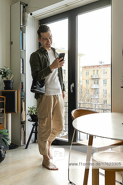 Happy young man using smart phone by window at home