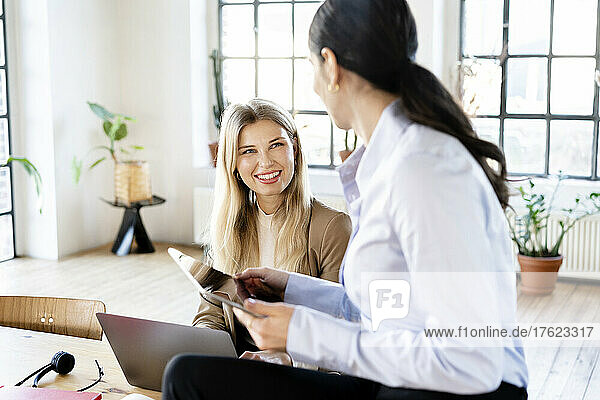 Happy blond businesswoman looking at colleague holding tablet PC