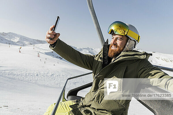 Smiling young man taking selfie through mobile phone sitting on ski lift