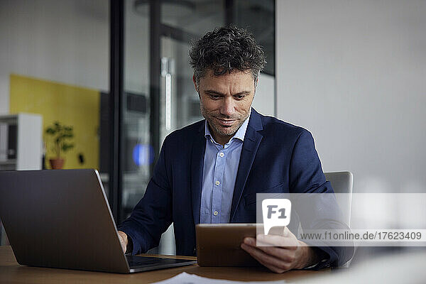 Smiling businessman using tablet PC sitting with laptop at desk in office