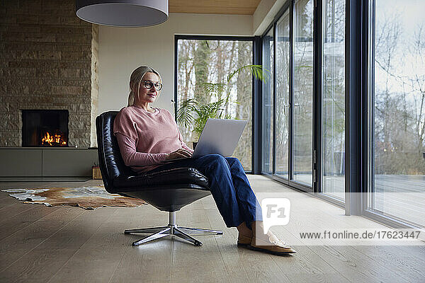 Blonde Frau mit Brille und Laptop sitzt auf einem Stuhl im Wohnzimmer