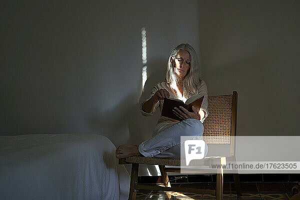 Woman reading book sitting on chair by bed at home