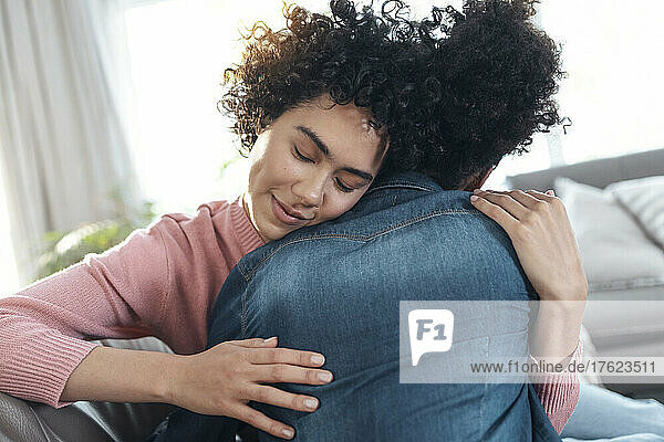 Woman with eyes closed embracing girlfriend sitting on sofa at home