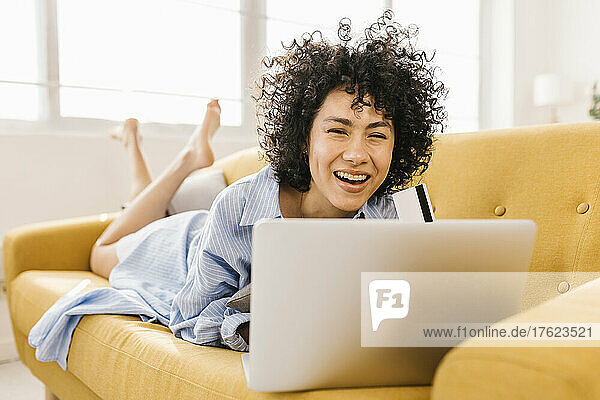 Cheerful young woman with credit card and laptop lying on sofa in living room at home