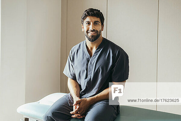 Lächelnder junger Physiotherapeut sitzt auf einer Trage in der medizinischen Klinik