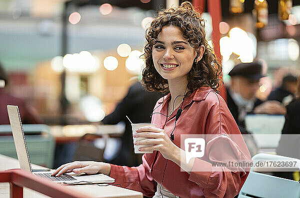 Smiling freelancer with disposable coffee cup and laptop in cafe