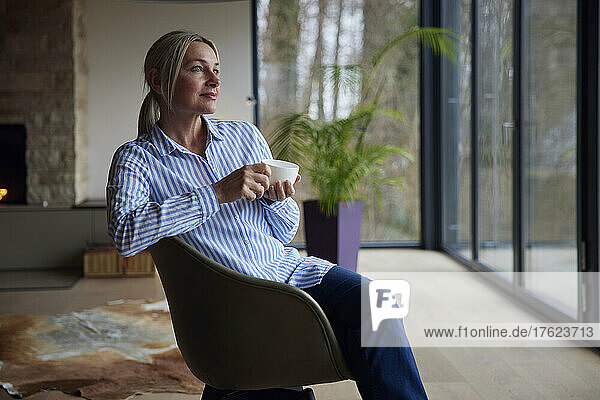 Thoughtful blond woman holding coffee cup sitting on chair at home