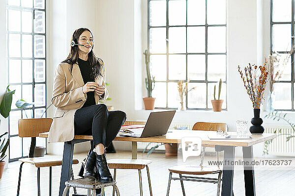 Smiling businesswoman talking through headset sitting on table at home office