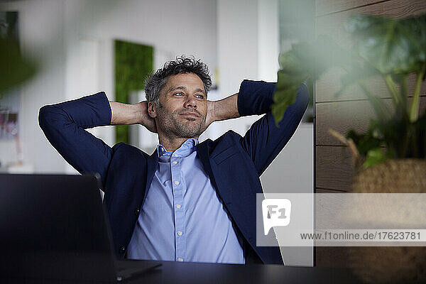 Smiling thoughtful businessman with hands behind head at work place